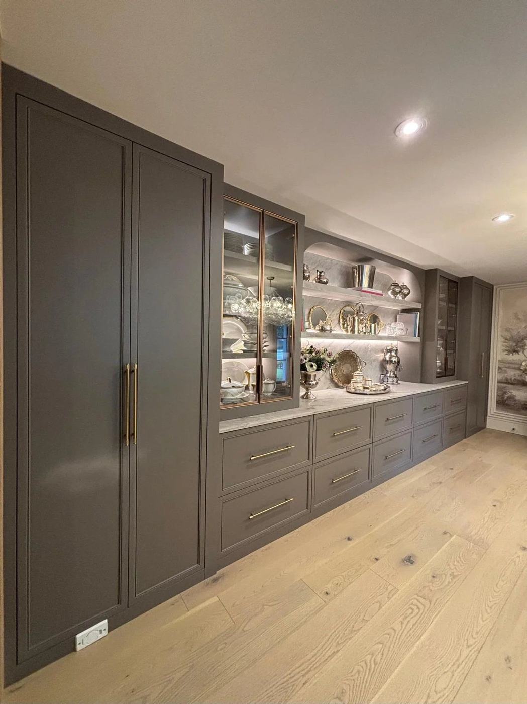 Wall of custom built-in cabinets in taupe with glass display doors, lit open shelving, a marble-look counter and brass hardware, by NewGen Renovations Vaughan