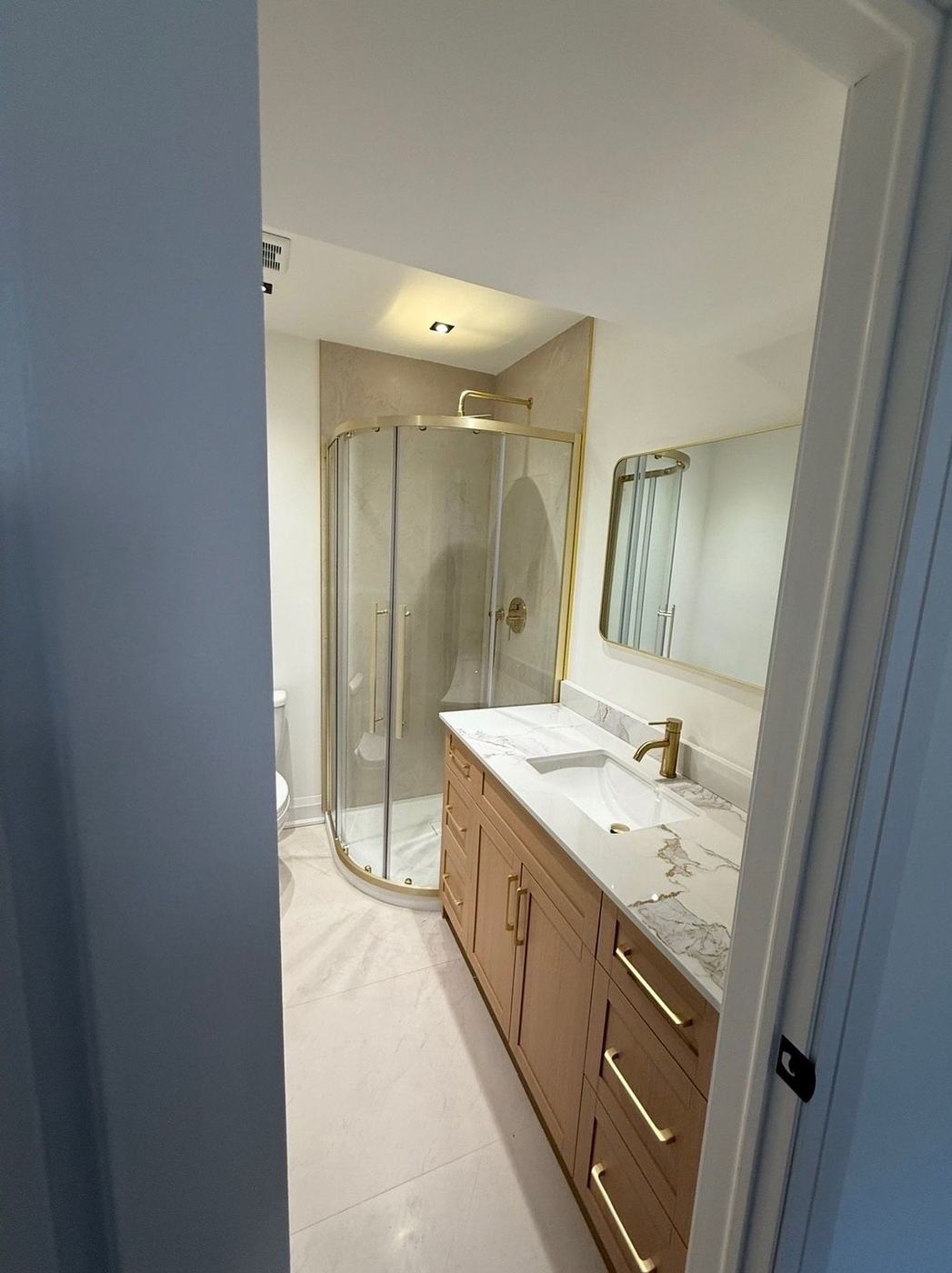 Bathroom with a curved glass corner shower, brushed gold fixtures and mirror, an oak shaker vanity with gold pulls and a veined quartz top by NewGen Renovations Vaughan